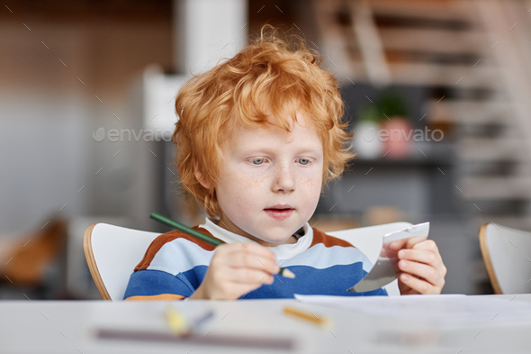Cute diligent gingerhaired learner of nursery school with crayon of dark green color drawing by desk at lesson during individual work